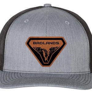 May include: A grey trucker cap with a black mesh back and a curved peak. The front displays a brown leather patch with the word "BADLANDS" and a black mountain graphic.