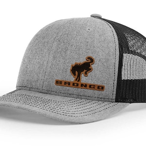 May include: A gray and black trucker hat with a brown leather patch featuring a black silhouette of a horse and the word "BRONCO".