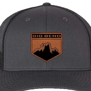 May include: A charcoal gray trucker hat with a black mesh back and a curved brim. The front features a brown leather patch with the words "BIG BEND" above a black mountain range graphic.