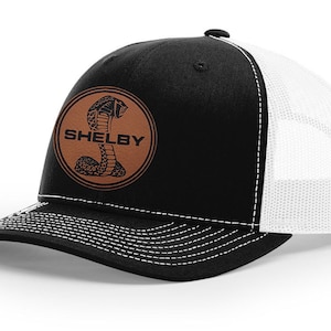 Peut inclure: Casquette de camionneur noire et blanche avec un patch en cuir marron représentant un serpent et le mot "SHELBY".
