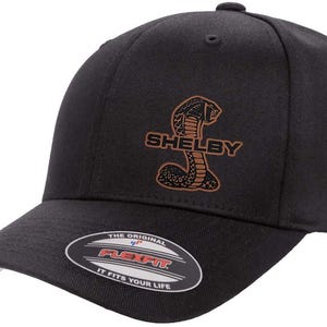 Peut inclure: Casquette de baseball noire Flexfit avec un logo cobra marron et le mot "SHELBY". La casquette a une visière incurvée avec un dessous blanc et une étiquette Flexfit.