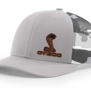 May include: A gray and black camouflage trucker hat with a brown leather patch featuring a snake and the text "GT 500".