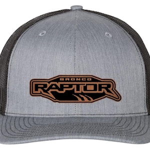 May include: A gray trucker hat with a black mesh back and a curved brim. The front features a brown and black patch with the word "RAPTOR" and the word "BRONCO" above it.