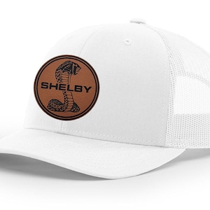 Peut inclure: Casquette de camionneur blanche avec un patch en cuir marron représentant un cobra noir et le mot "SHELBY" en lettres noires.