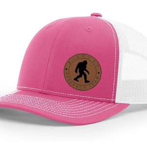 May include: Pink and white trucker hat with a brown leather patch that says "Bigfoot Research Team" with a silhouette of Bigfoot walking.