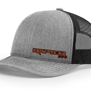 May include: A gray and black trucker hat with a brown embroidered logo that says "RAPTOR SVT".