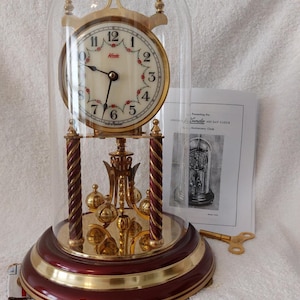 May include: A gold-toned Kundo 40-day anniversary clock with a glass dome. The clock has a white face with black Roman numeral hour markers and a red floral design. The clock is sitting on a red wooden base with a gold trim.