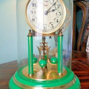 Può includere: Un orologio vintage verde e dorato con una cupola di vetro. Il quadrante dell'orologio ha lancette nere e numeri romani. L'orologio è su una base verde.
