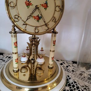 May include: A gold-toned Kundo brand mantle clock with a white face and floral accents. The clock has a pendulum and a glass dome.