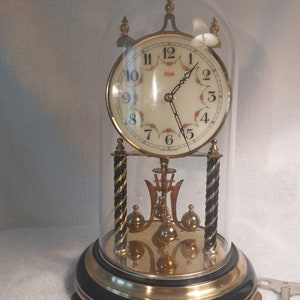May include: A vintage gold-toned clock with a white face and black Roman numeral markings. The clock is encased in a glass dome with a black base and gold trim. The clock has a pendulum and is decorated with small red flowers.
