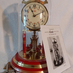 May include: A gold-toned Kundo 400-day clock with a glass dome, red base, and gold accents. The clock face has a white background with black Roman numerals and a black hand. The clock is on a white surface with a paper instruction sheet.