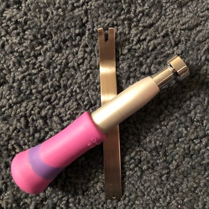 May include: A metal upholstery staple remover tool with a flat, forked tip and a silver metal shaft, paired with a separate tool featuring a pink and purple ergonomic handle and a silver metal cylindrical head with a flat, circular top surface.