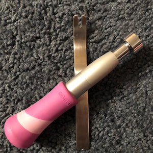 May include: A metal upholstery tool with a forked end and a handle featuring a pink and light pink diagonal striped grip. The handle has the text PAT PEND embossed on the side. The tool is positioned vertically with a silver metallic finish.