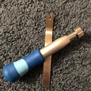 May include: A metal pry tool with a forked end and a separate tool featuring a silver metallic shaft, a cylindrical head, and a handle with alternating bands of dark blue and light blue rubberized material, positioned in a crossed arrangement.