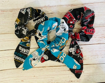 Puede incluir: Tres coleteros de tela con cintas largas. Uno es negro con logotipos y texto de los Jacksonville Jaguars. Uno es turquesa con logotipos y texto de los Miami Dolphins. Uno es negro con logotipos de los Tampa Bay Buccaneers y texto rojo y blanco.