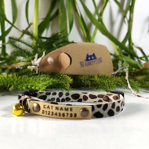 Puede incluir: Un collar de gato de estampado de leopardo marrón y blanco con una campanita dorada y una placa de nombre plateada que dice "CAT NAME 0123456789".