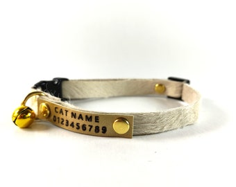 May include: A white faux fur cat collar with a gold metal buckle and a gold bell. The collar has a gold tag with the text "CAT NAME 0123456789" engraved on it.