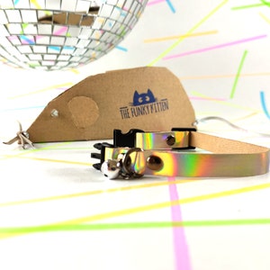 Op de afbeelding: Een holografische, iriserende kattenhalsband met een zilveren bel en een zwarte gesp. De halsband ligt op een witte achtergrond met kleurrijke lijnen. De tekst "The Funky Kitten" staat op een bruin kartonnen label.