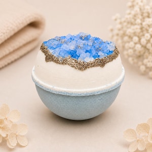 May include: A spherical bath bomb with a smooth white upper half and a light blue lower half. The top features a cluster of translucent blue crystals surrounded by a textured band of gold glitter, mimicking the appearance of a geode formation.