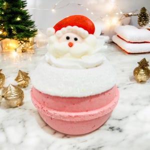 Puede incluir: Una bomba de baño festiva con forma de Papá Noel. Presenta un gorro rojo, barba blanca y base rosa. La parte superior está decorada con glaseado blanco y purpurina. Tema navideño.