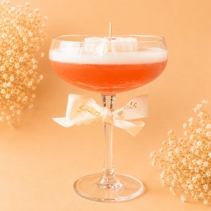 Può includere: Una candela in una coppa di vetro riempita di cera color corallo, sormontata da un accento di cera a forma di fiore bianco e uno stoppino centrale. Un nastro color crema con il testo dorato "Soak Nights" è legato attorno allo stelo del calice.