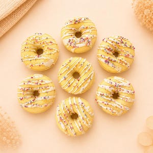 Pode incluir: Sete donuts amarelos redondos dispostos num padrão circular. Cada donut apresenta uma cobertura de glacê branco e é decorado com pequenos granulados coloridos. Os donuts são apresentados sobre uma superfície de fundo cor de pêssego.