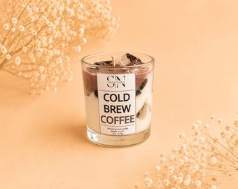 Może przedstawiać: Świeca w przezroczystym szklanym słoiku z warstwowym białym i brązowym woskiem. Etykieta zawiera napis SN Soaky Nights COLD BREW COFFEE, Hand poured candle, Made in U.K., 4oz / 120g. Na wierzchu znajdują się ciemne kawałki wosku przypominające kostki lodu.