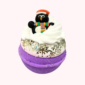 Puede incluir: Una bomba de baño festiva con una base morada, una capa intermedia rosa y dorada con purpurina y una parte superior blanca de crema batida. Un pingüino negro con un gorro rojo y una bufanda colorida está encima. Un divertido producto de baño temático navideño.