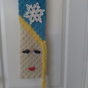 May include: A crocheted scarf with a blue and white snowflake pattern and a yellow braid. The scarf also features a crocheted face with blue eyes, red lips, and yellow hair.