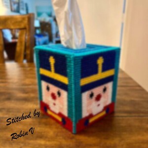 May include: A handmade tissue box cover featuring a soldier design. The box is primarily blue, with yellow and red accents. The soldier's face is pink with red details. The text "Stitched by Robin V" is on the front.
