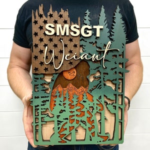 Puede incluir: Un letrero de madera con un recorte de un Bigfoot caminando por un bosque. El letrero está pintado en tonos de marrón, verde y blanco. El texto "SMSGT Weiant" se muestra en el letrero.