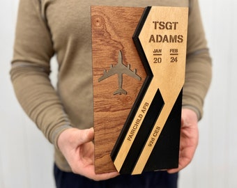 Puede incluir: Una placa de madera con un diseño geométrico en negro y marrón. La placa presenta un recorte de un avión, el texto "TSGT ADAMS", "JAN 20", "FEB 24", "FAIRCHILD AFB", y "92d CES".