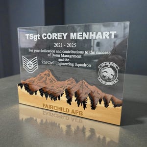 Può includere: Una targa in acrilico trasparente e legno con testo inciso e una scena di montagna. Il testo recita "TSgt COREY MENHART 2021-2025" e i dettagli del servizio. La base presenta una catena montuosa scolpita e le parole "FAIRCHILD AFB".