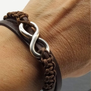 Puede incluir: Un brazalete de cuero marrón con un dije de plata en forma de símbolo de infinito. El brazalete está trenzado y tiene un aspecto de capas.