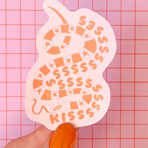 May include: A die-cut sticker featuring a light pink background with a coiled snake graphic. The snake has alternating peach-coloured and light pink segments decorated with white stars. The text "KISSSS" is printed in peach at the bottom.