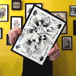 May include: Black and white framed artwork featuring a stylized cartoon character with a star-shaped face, surrounded by barbed wire and radiating lines. The artwork is held against a yellow wall with other framed pieces.