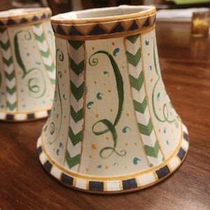 May include: Two decorative lampshades with a cream-colored background. The shades feature green, blue, and yellow accents, including chevron patterns and whimsical swirls. The rims are trimmed with a navy blue and gold border.