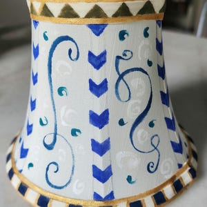 May include: A small, white lampshade with a decorative design. The shade features blue chevron and swirl patterns, with teal accents. Gold trim accents the top and bottom edges. The lampshade is approximately 5 inches tall.
