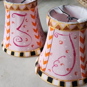 May include: Two small, bell-shaped lamp shades featuring a light pink fabric base with hand-painted magenta swirls, orange chevron vertical stripes, and a gold-trimmed top border. The bottom edge is finished with a black and white striped trim.