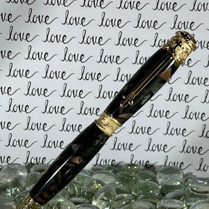Op de afbeelding: Een zwart-gouden pen met sierlijke gouden accenten. De pen heeft een gemarmerd patroon en ligt op een bed van heldere glazen stenen. De achtergrond toont het woord "liefde" herhaald in een handgeschreven stijl.