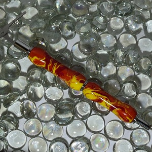 May include: A close-up of a red and yellow swirl pen with a silver tip and a silver band. The pen is lying on a bed of clear glass beads.