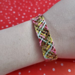 Puede incluir: Un brazalete de amistad colorido con un patrón de chevron en tonos de marrón, rosa, amarillo, verde y blanco.