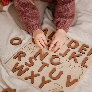 Op de afbeelding: Een kind speelt met een houten alfabetpuzzel. De puzzel heeft alle letters van het alfabet in afzonderlijke stukken. Het kind houdt de letter C in zijn hand.
