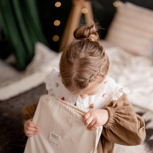 Op de afbeelding: Een jong meisje met haar haar in een knot zit op een bed en opent een witte trekkoordtas. De tas heeft een klein label aan de voorkant met de tekst "remarkable".