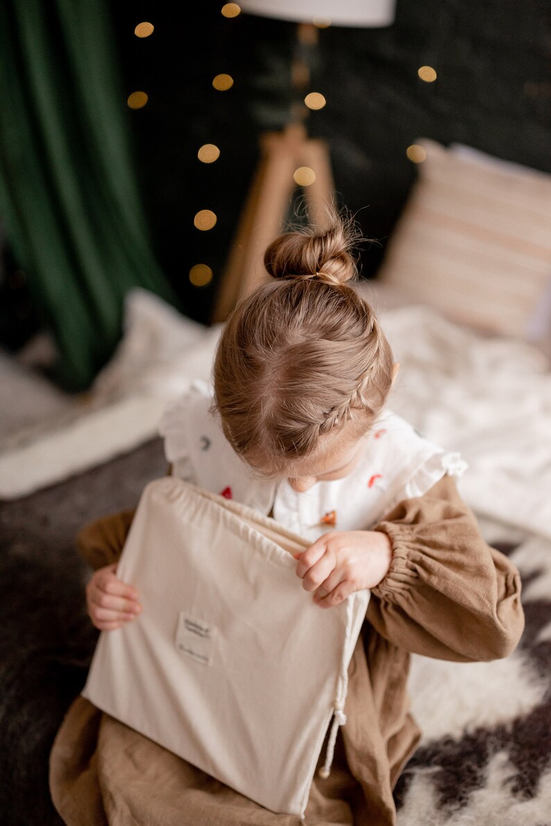 Op de afbeelding: Een jong meisje met haar haar in een knot zit op een bed en opent een witte trekkoordtas. De tas heeft een klein label aan de voorkant met de tekst "remarkable".