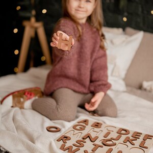 Op de afbeelding: Een jong meisje houdt de letter "H" omhoog van een houten alfabetpuzzel. De puzzel ligt op een wit deken met de andere letters zichtbaar.