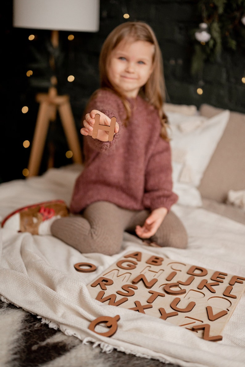 Op de afbeelding: Een jong meisje houdt de letter "H" omhoog van een houten alfabetpuzzel. De puzzel ligt op een wit deken met de andere letters zichtbaar.