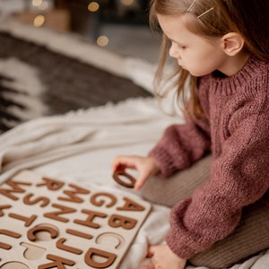 Op de afbeelding: Een kind zit op een wit deken en speelt met een houten alfabetpuzzel. De puzzel heeft bruine letters en ligt op het deken. Het kind draagt een roze trui.