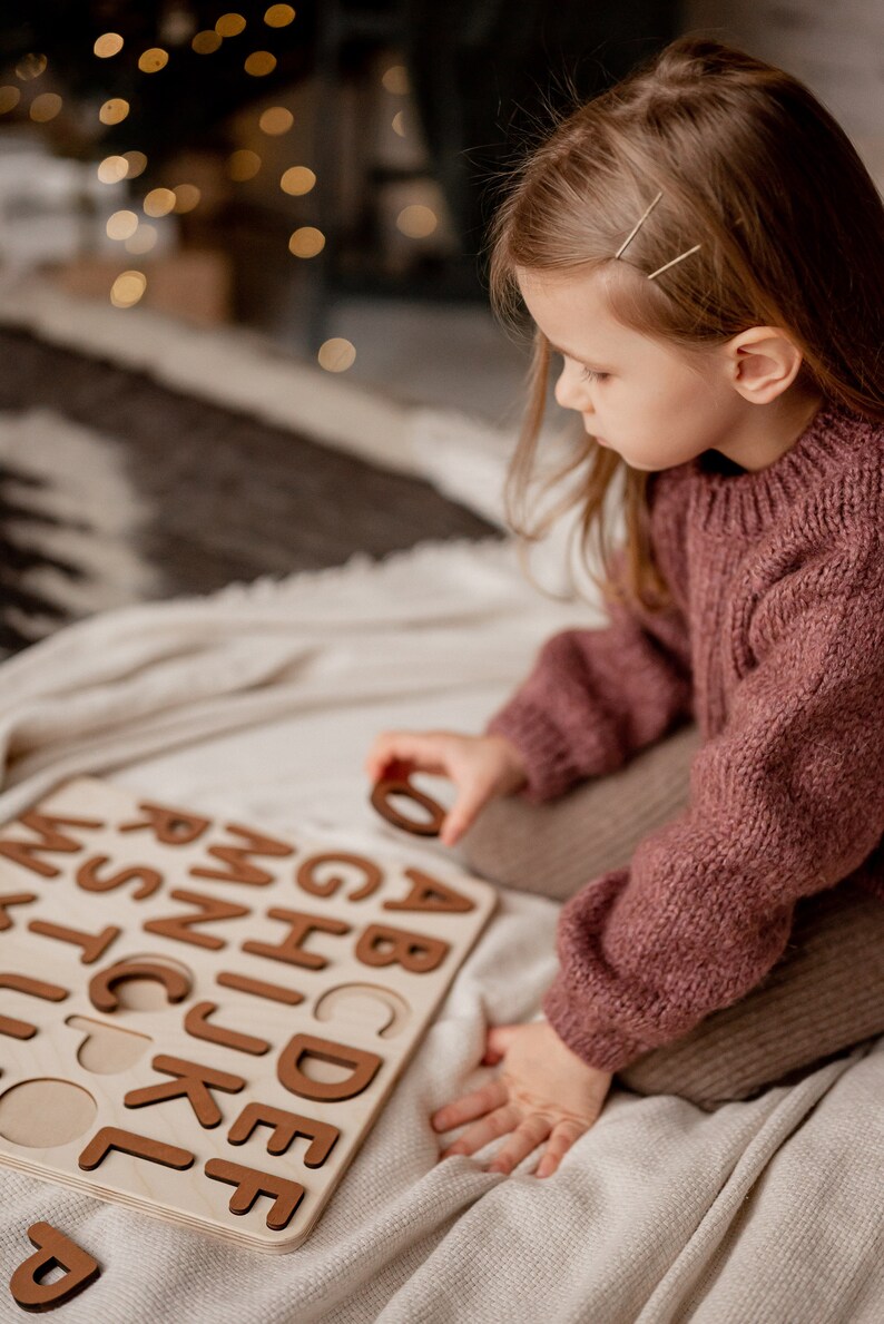 Op de afbeelding: Een kind zit op een wit deken en speelt met een houten alfabetpuzzel. De puzzel heeft bruine letters en ligt op het deken. Het kind draagt een roze trui.