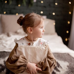 Op de afbeelding: Een jong meisje in een bruine jurk zit op een bed en houdt een wit kussen vast. Ze kijkt naar de zijkant en de achtergrond is wazig.